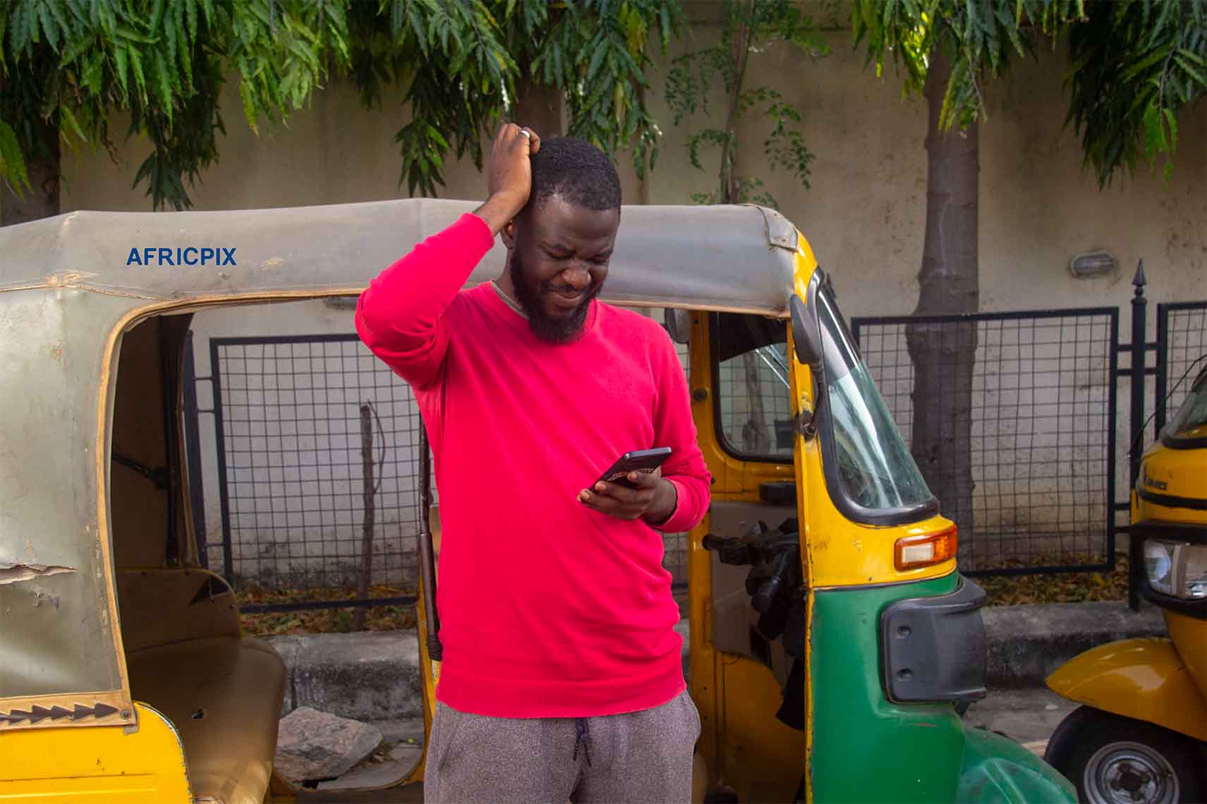 A confuse African tricycle rider standing with his tricycle, Keke, Maruwa behind him, happy, looking confused, hand on his head, while his holding his phone